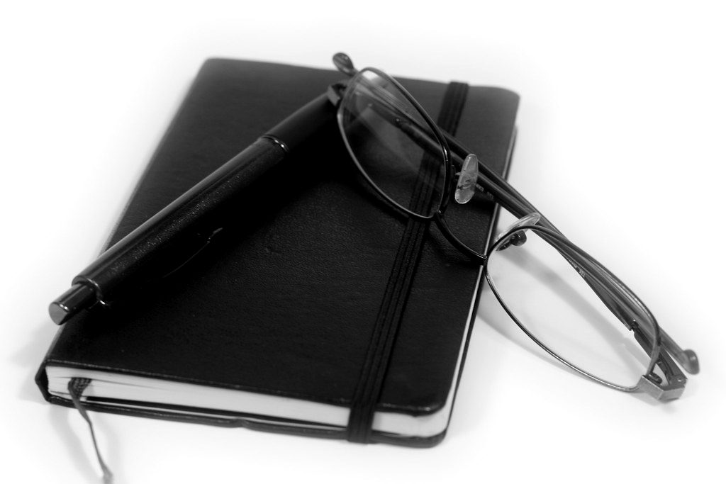 A black notebook, pen and reading glasses
