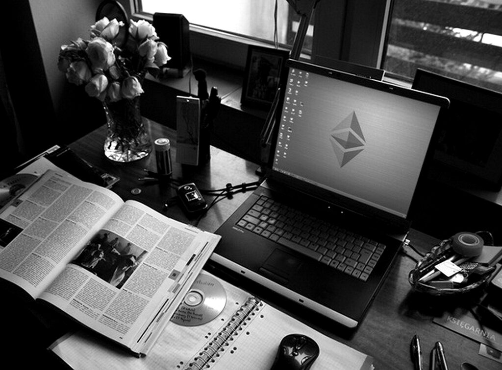 A laptop showing the Ethereum logo on a desk beside a newspaper and roses